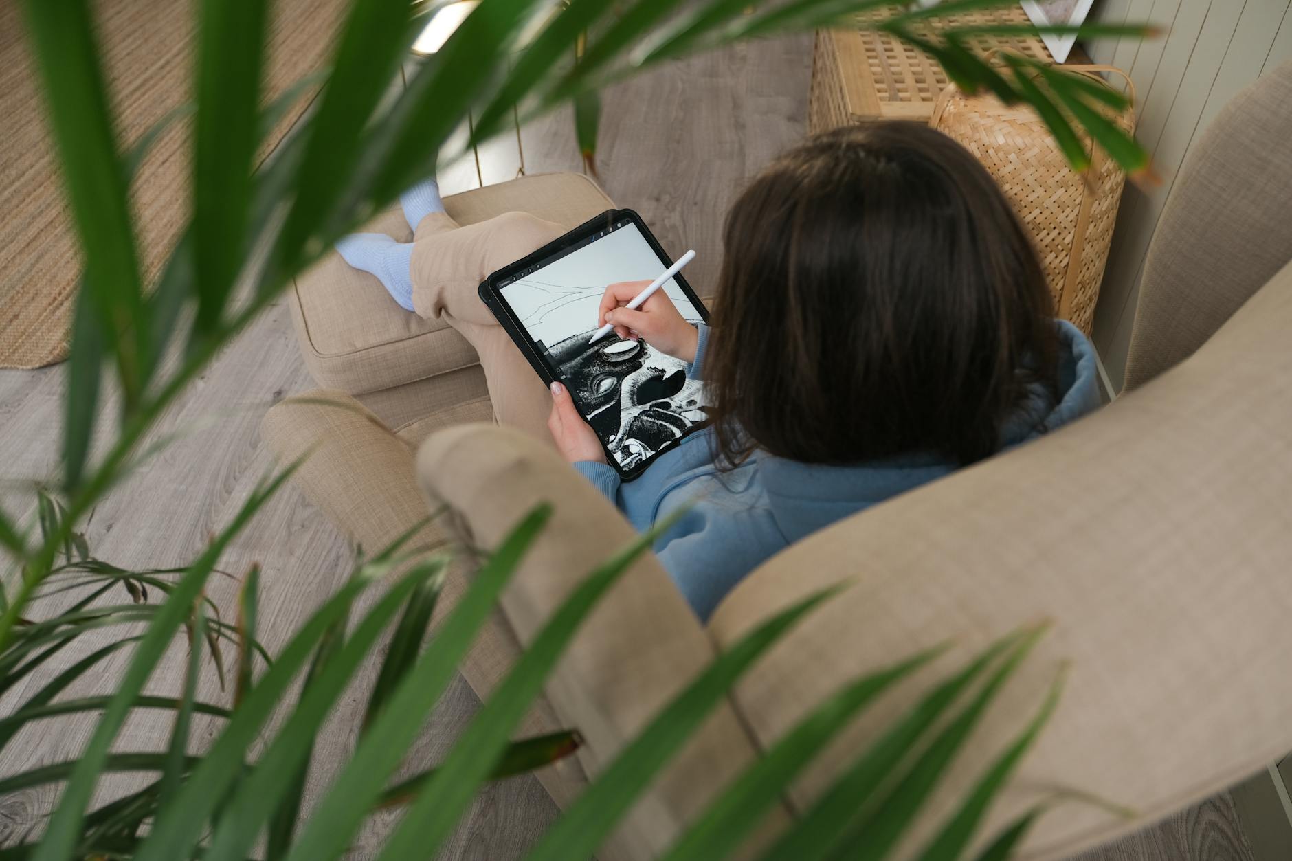 A person drawing on a digital tablet while lounging on a sofa, surrounded by plant leaves.
