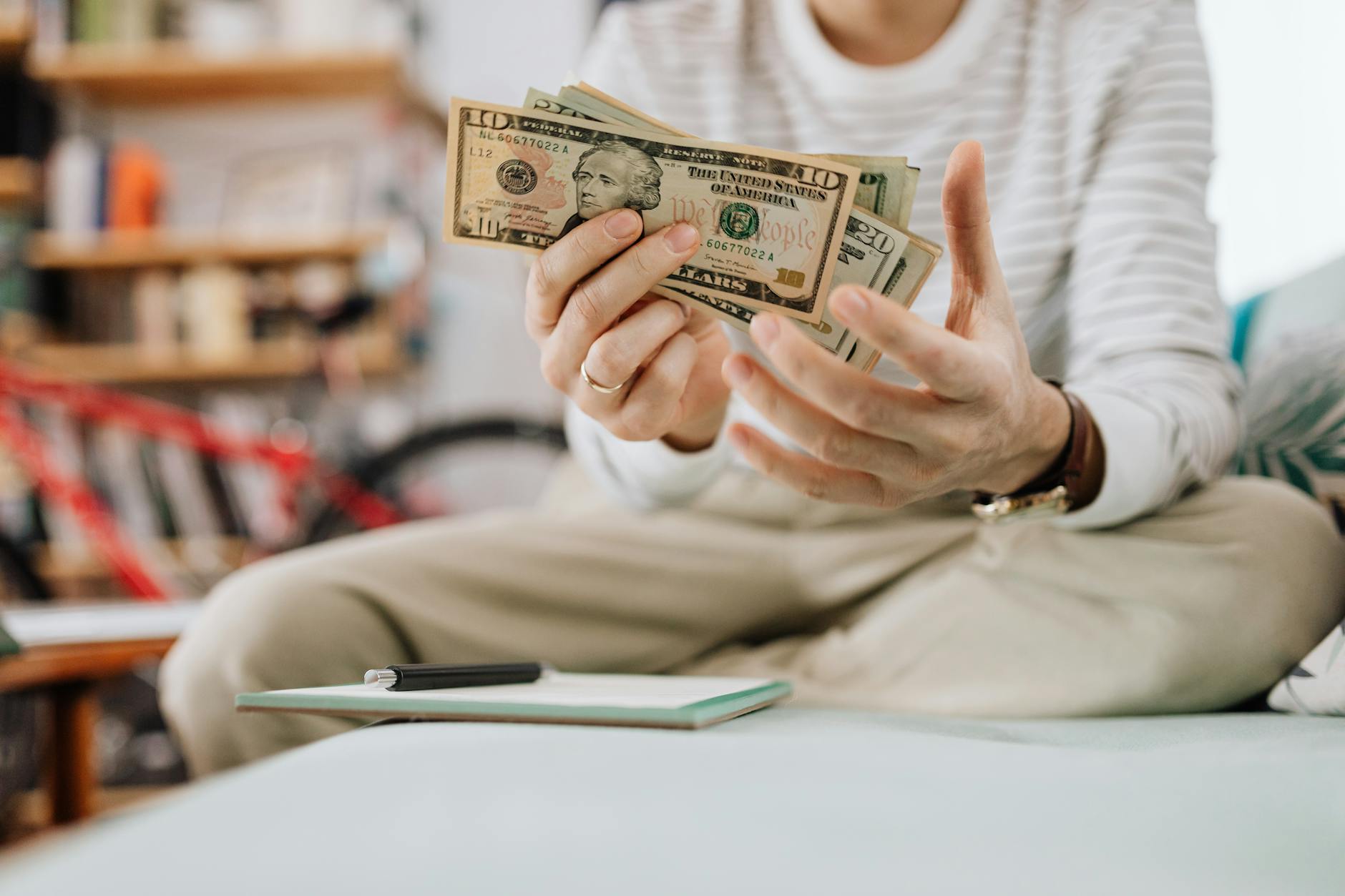 Man wearing casual clothes counts dollar bills while sitting on a sofa indoors.